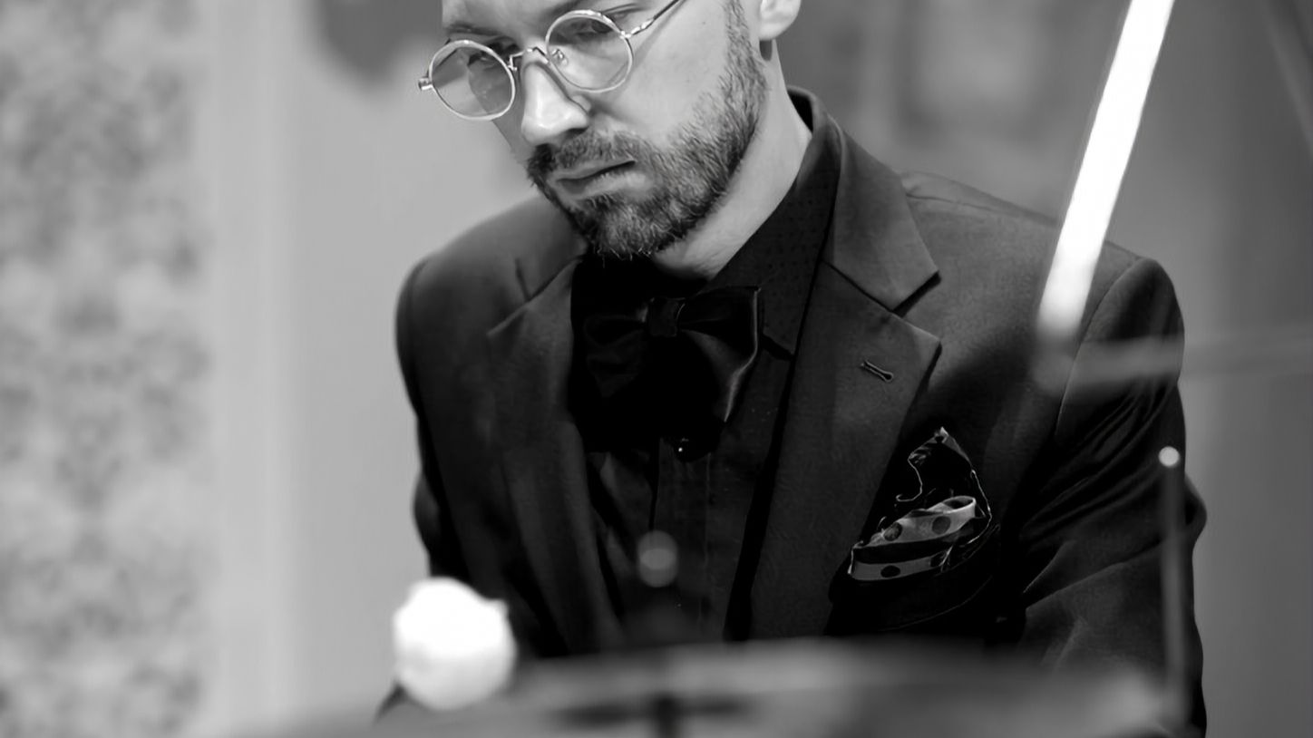 Black-and-white close-up of Łukasz Kurzydło in round glasses and a bow tie, playing with a mallet