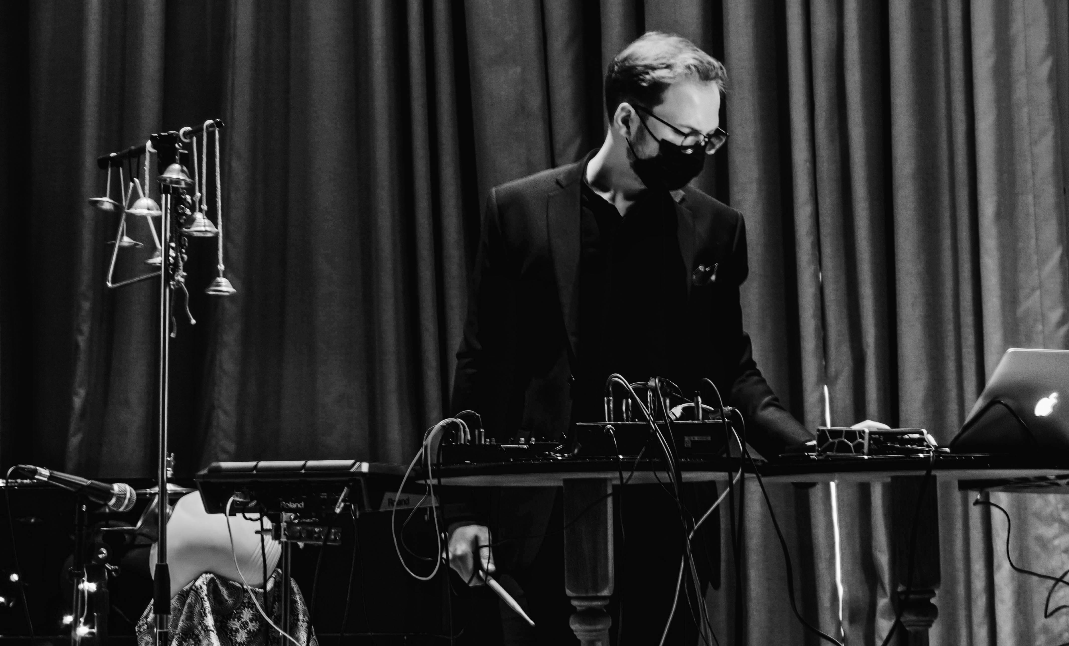 Black-and-white photo of Łukasz Kurzydło in a face mask at a table of synthesizers and a laptop on stage, with hanging bells beside him