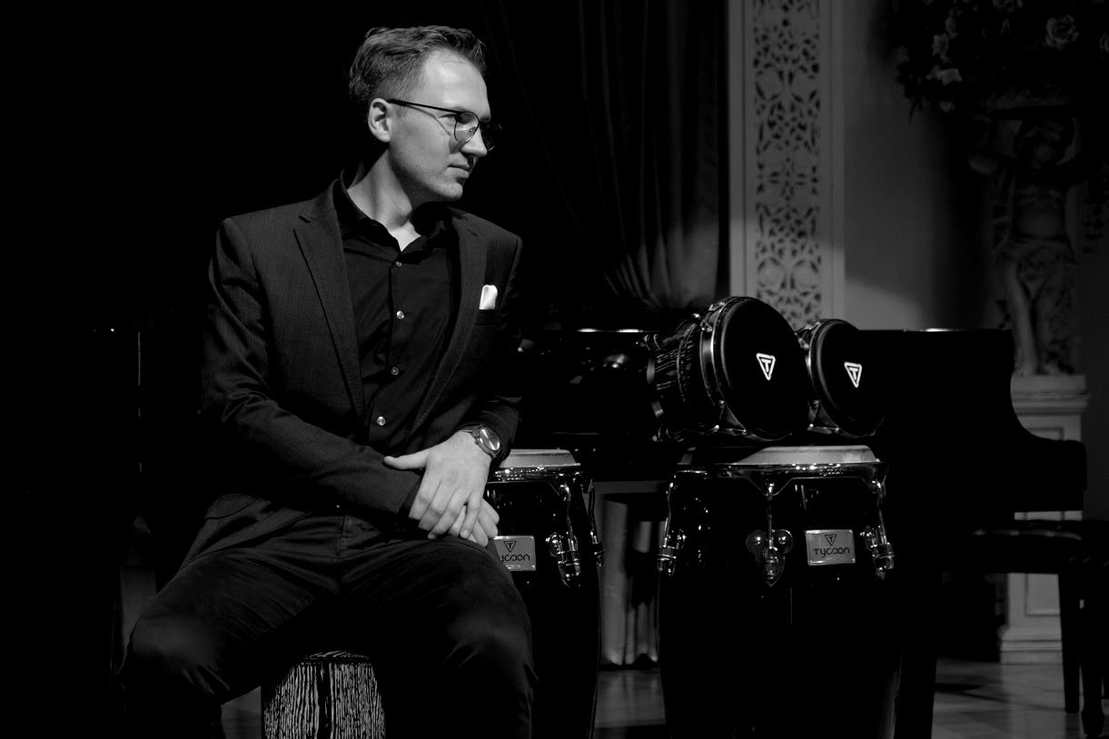 Black-and-white photo of Łukasz Kurzydło in a suit, seated beside a pair of congas on a concert stage